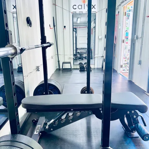 Inside view of Calyx Fitness private training facility showing an adjustable bench press setup with barbell, weight plates, and the Calyx logo visible in the background. The intimate, private gym environment offers personalized training space.